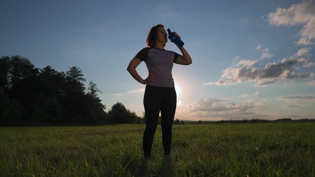 A woman stands in a field, holding a sports bottle while enjoying the outdoors. The woman gazes at the horizon, showcasing her involvement in sport.