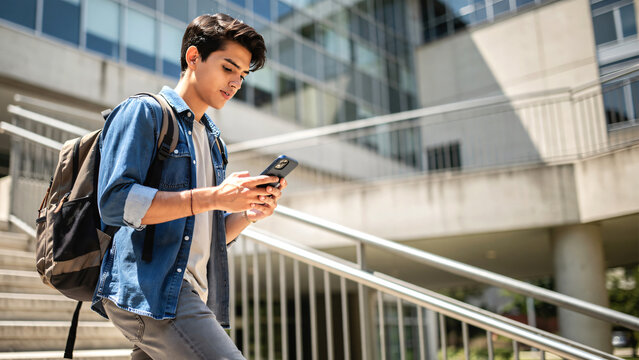 Young male university student using a smartphone while walking down stairs on campus, focused on screen with natural casual expression. 