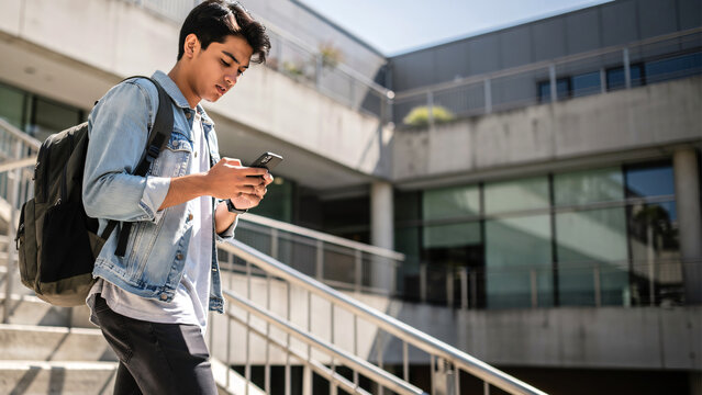Young male university student using a smartphone while walking down stairs on campus, focused on screen with natural casual expression. 