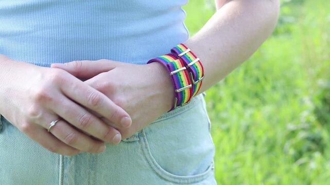 Woman in light blue top with rainbow LGBT bracelet on wrist against blurred green background. Close-up of young girl's hands with bright rainbow textile pride strap, tolerance and summer concept