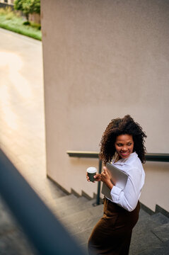 African American Woman Carrying Laptop And Coffee Walking Up Stairs Outside Office Building, Smiling Confidently