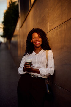 African American Business Woman Smiling With Coffee Against City Wall At Golden Hour