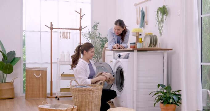 Asian LGBTQ female couple transfers mixed laundry from basket into washing machine, lifting soft clothing and smiling during weekend home routine, shared housework, support, teamwork and affection
