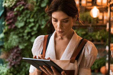 Fototapeta premium Woman Using Digital Tablet In Garden Center With Potted Plants And Warm Lighting Background