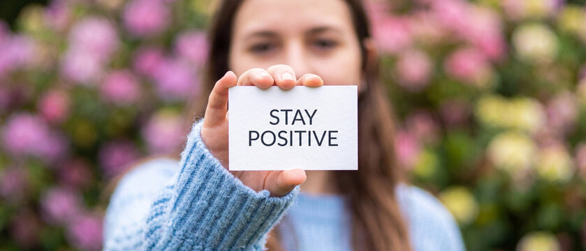 Young woman holding a sign with the text stay positive in front of a blurred floral background