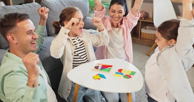 Family parents children girls cheering victory at home. They are playing a puzzle game at a small table, celebrating with raised hands and smiles. Concept is family win and play together.