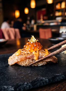 Close-up of premium aburi sushi nigiri topped with salmon roe, sauce and edible gold leaf held by chopsticks on a black slate plate with a moody restaurant background.