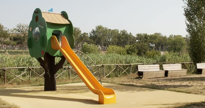 A colorful children's playground slide structure shaped like a green treehouse with a yellow slide and climbing arch, set on a padded play surface outdoors surrounded by green trees under a sunny sky.
