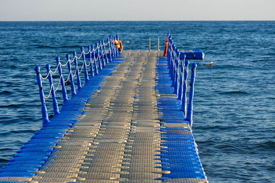 Floating pontoon jetty with blue safety railing built on water surface of the Mediterranean Sea at a coastal holiday resort on a sunny day.