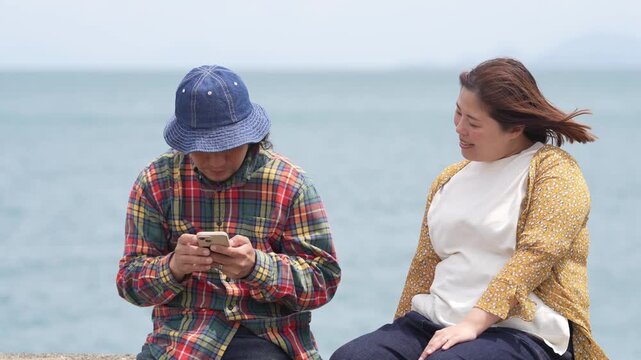 Three people sit by the sea, relaxed and smiling. One person wears a bucket hat and checks their phone. The others enjoy the ocean breeze and each other's company. Perfect for lifestyle, travel