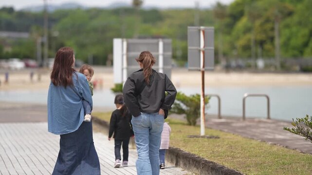 Two women and children stroll along a paved path. Lush greenery frames a tranquil waterfront. They move casually, suggesting leisurely enjoyment. Perfect for family outings or serene park scenes