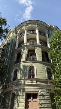 Historic green building facade with white columns, elegant caryatid statues supporting curved wrought-iron balconies, and arched windows framed by green trees under a blue summer sky.