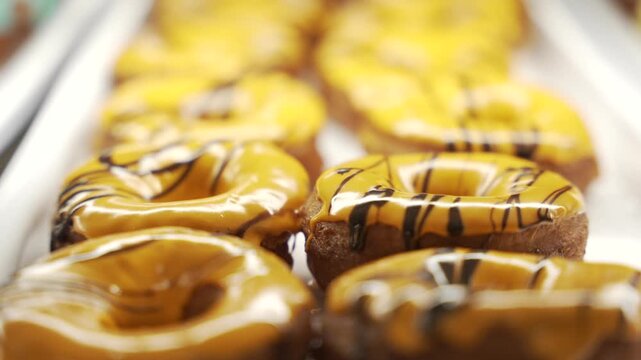 Yellow glazed ring donuts with chocolate drizzle sit in rows on a metal bakery tray, shot in soft focus for a tempting dessert closeup.
