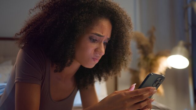 Frustrated African American woman using mobile phone losing bet online failure in bed at night disappointed ethnic girl female scrolling smartphone lose internet betting user upset lying in bedroom