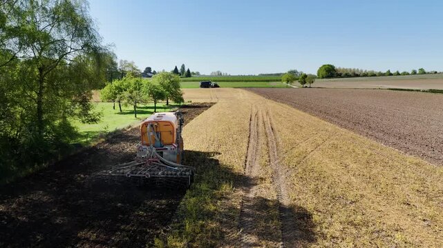 Aerial drone tracking shot of a yellow self-propelled slurry manure injector fertilizing a sunny agricultural field.