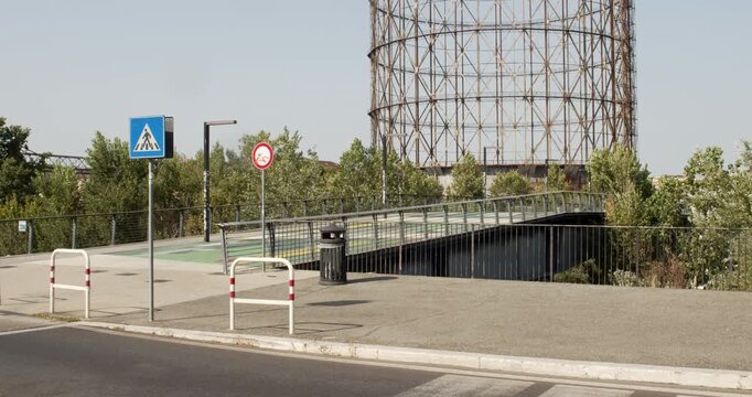 A pedestrian bridge with glass railings leading towards green trees, with road signs, street barriers, and a large metal gasometer structure looming in the background. It's located in Rome, Italy.
