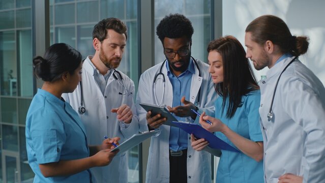 Focused multiethnic five doctors with digital tablet working talking deciding medicine diagnosis in clinic hallway. Multiracial nurses practitioners therapists surgeons talk team work cooperative