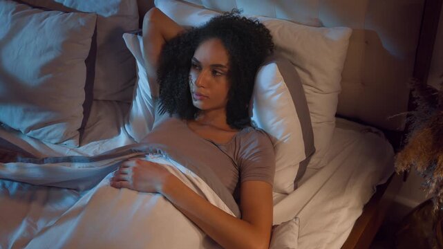 Nervous African American woman suffering with insomnia napping sleeping difficulties in bed at night girl female awake by noisy neighbors female trying to sleep uncomfortable position lying in bedroom