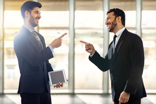 Corporate employees in formal business suit pointing forefingers and laughing with one of them holding digital tab.