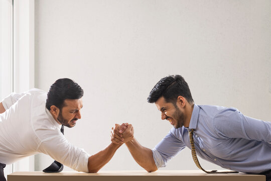 Corporate employees in formal clothing engaged in an arm wrestling match with one of them about to lose.