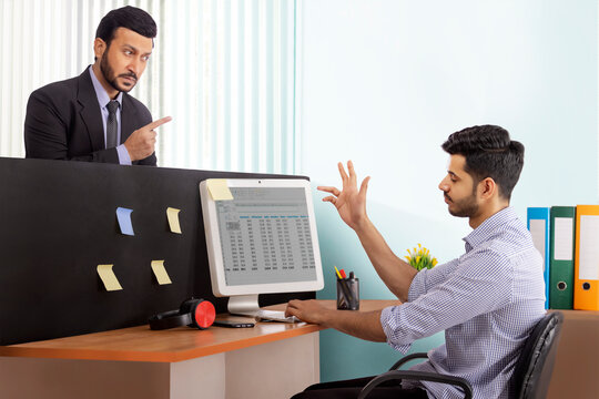 An angry boss in formal business suit scolding junior corporate employee at workdesk.