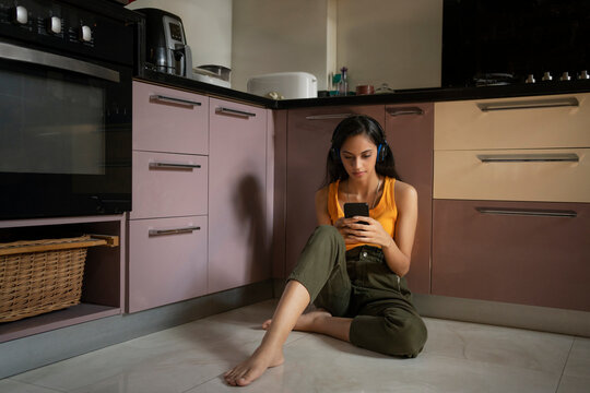 A young woman sitting in her kitchen listening to music on headphones with mobile in hand.