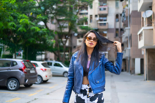 A young woman standing outside flats wearing a crop top and leather jacket playing with her hair.