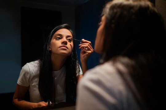 A young woman applying mascara with a wand sitting in front of mirror in a room.