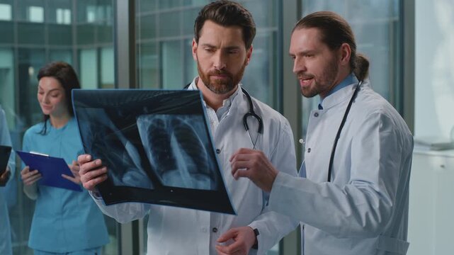 Two professionals Caucasian doctors analyzing x-ray talking serious deciding diagnosis in modern clinic. Colleagues males men medical surgeons therapists talk discussing lungs scan image medicine help
