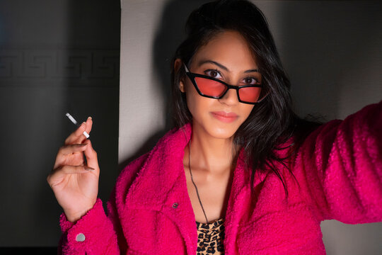 A young stylish woman wearing eyeglasses holding a cigarette in hand.