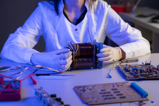 A person in a lab coat tests an electrical coil with tools in a research lab setting at night