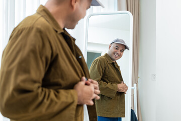 Fototapeta premium Smiling Asian man buttoning his casual brown jacket in front of a mirror. Concept of morning routine preparation, trendy street fashion and everyday confidence.