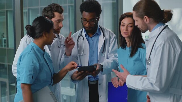 Group of five multiethnic medical workers doctors looking in digital tablet talking concentrated working discussing web medicine health care in modern clinic. Team of diverse surgeons talk partnership