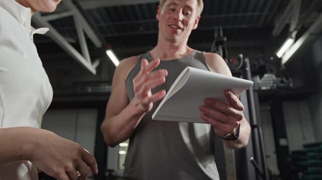 Low angle shot of young male fitness trainer holding notepad and explaining workout plan to female client in gym. Ideal for promoting personal training services