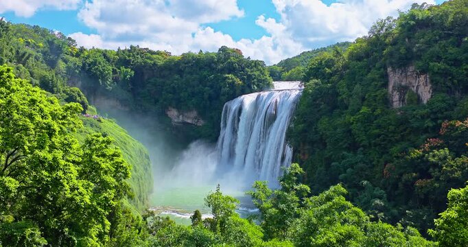 Huangguoshu waterfall in Anshun, Guizhou, China. Beautiful view of a large waterfall surrounded by lush green forests and a turquoise plunge pool under a bright sky.