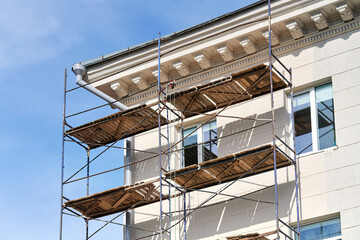 Industrial scaffolding setup for house painting and plastering works. Multi level scaffolding with wooden planks erected against classical white stucco facade of building under renovation on sunny day © Tricky Shark