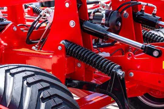 Agricultural machinery hydraulic cylinder and chassis tension spring. Modern farm machine pressure system component on metal frame with transport vehicle low pressure tire close up details.