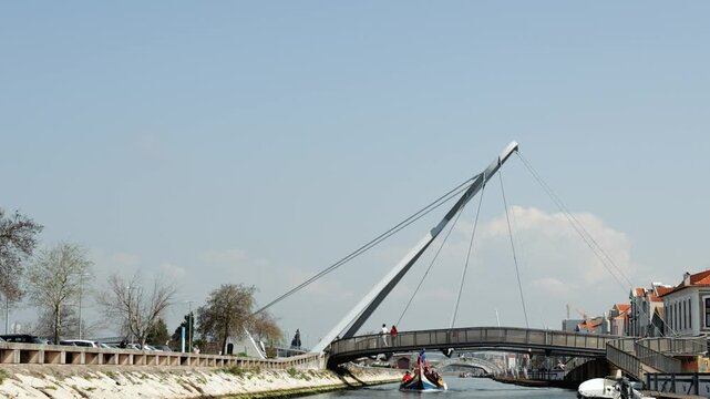 High-quality water-level video following a traditional colorful Moliceiro boat filled with tourists sailing through the Ria de Aveiro canal. Boat moves towards a modern bridge on a bright sunny day