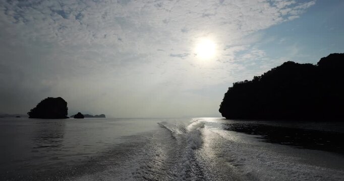 Slow motion view looking back from fast motorboat returning from geopark tour at Langkawi. Silhouetted karst islets rise at calm bay, sun shines through light clouds, wake from boat on water