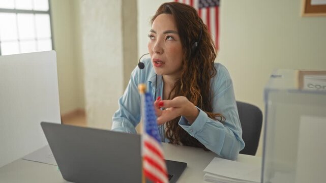 Woman with headset gestures hand toward laptop and small american flag at a polling table inside a civic building; civic duty focus.