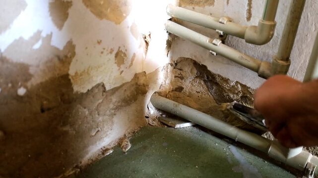 Worker carefully applies liquid primer to an old concrete wall around plastic pipes in a room corner. This video close-up details the surface preparation stage, moisture protection, and home renovatio