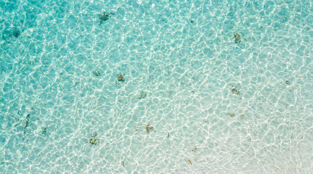 Clear turquoise water with gentle ripples and scattered small rocks on a sandy seabed, viewed from above.