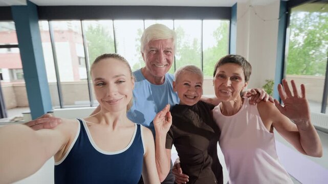 Portrait of Caucasian young adult female instructor and group of Caucasian senior people smiling and waving at camera in fitness studio. Footage represents active aging and wellness programs
