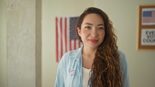 Woman smiling with an i voted sticker on her shirt and visible face, standing in a polling building with american flag and framed sign behind her; civic pride.