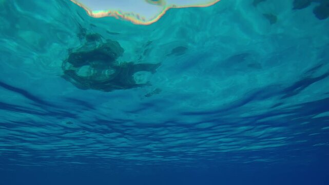 Ripples on the surface of blue water in calm weather with sandy bottom and corals reflected in it. Natural underwater background with water surface in daylight