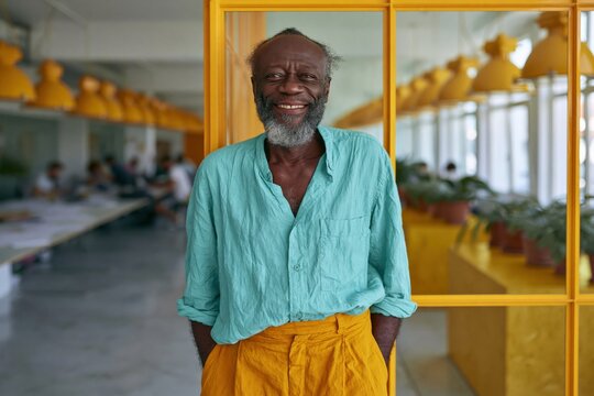 Elder man smiling in modern bright office interior wearing teal shirt and mustard trousers for corporate portrait and workspace banner