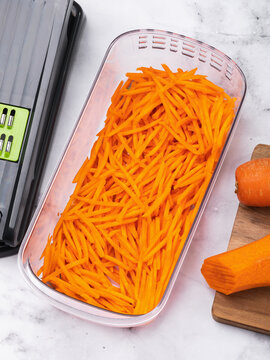 Fresh grated carrot julienne in open plastic container with vegetable slicer on marble table top view