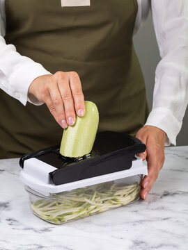 Woman grating fresh zucchini into julienne strips on manual mandoline slicer with container on marble table