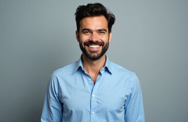 Obraz premium Happy bearded man smiles at camera. Young guy wears blue shirt in studio light. Professional male model looks confident with positive facial expression. Attractive person stands against simple grey