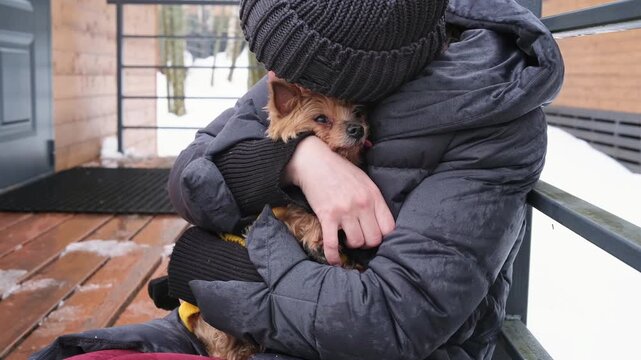 woman in winter jacket holds small dog shivering from cold on wooden deck. caring owner protects dog from cold winter weather. small dog needs warmth in cold season